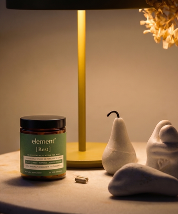 A jar of element3 [Rest] dietary supplement capsules sits on a tabletop next to a gold lamp, with decorative white ceramic pears and stones in the background.