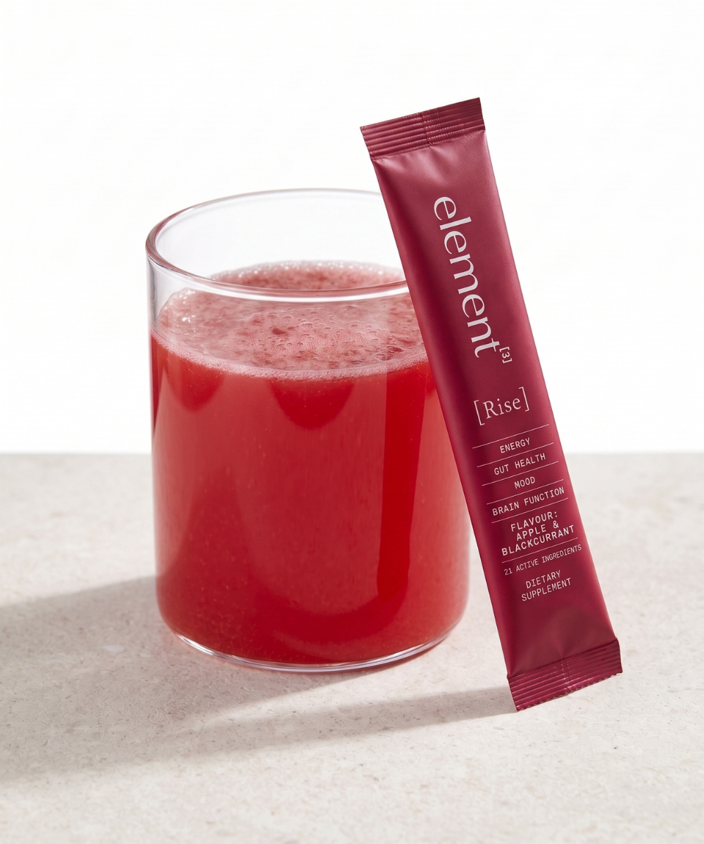 A glass of bright red beverage with a single Element3 [Rise] energy gut health sachet standing beside it on a light neutral surface.